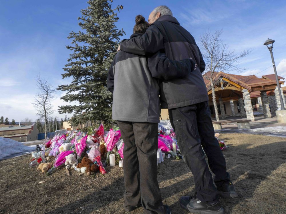 Two people wearing dark winter jackets embrace while standing before a memorial display of flowers laid at the foot of a tree on a bright winter day.