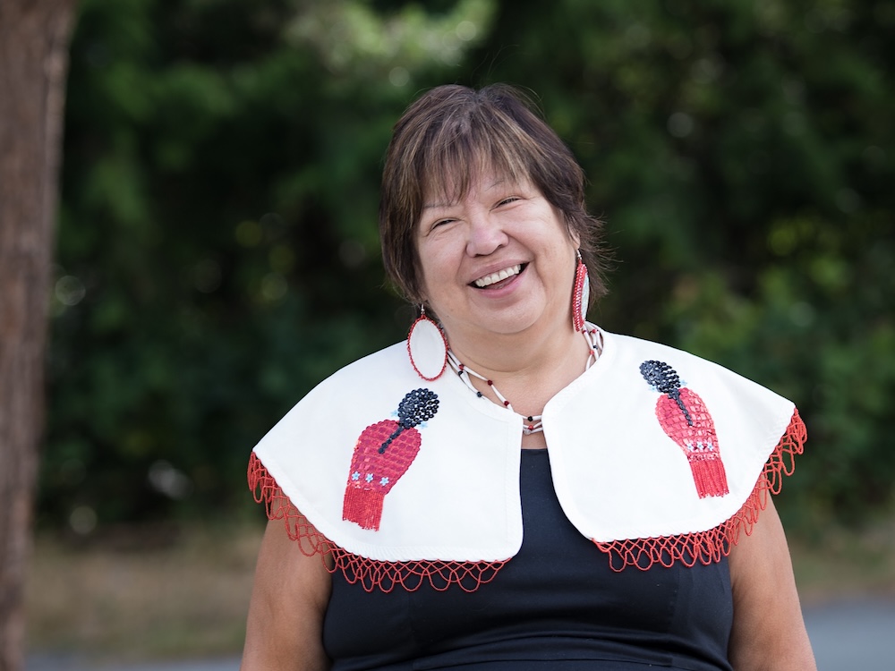 Judith Sayers, an Indigenous woman with short brown hair and medium-light skin tone, smiles at the camera. She is wearing an embroidered shoulder covering and large round earrings.