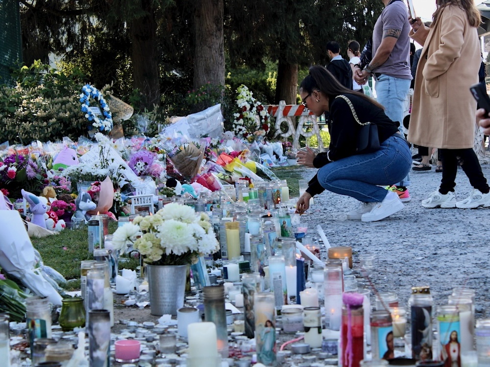 A woman wearing a black sweater and jeans crouches down in front of a pile of flowers and stuffed animals to light a candle. In front of the pile are numerous lit candles.