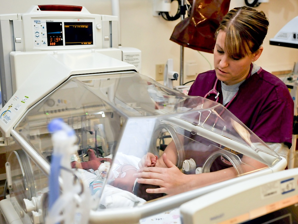 A woman in burgundy hospital scrubs with a stethoscope reaches into an incubator and holds an infant&rsquo;s hand.