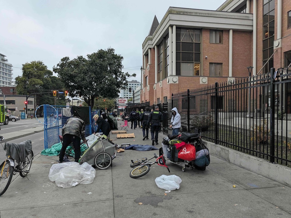 People who are apparently homeless and their possessions on a sidewalk as a group of police and bylaw officers walk past.