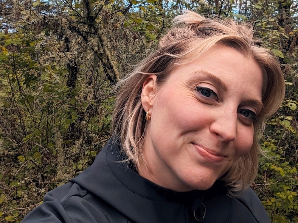 Laura Gover wears a black sweater and she has short wavy blond hair. She is smiling at the camera against a wooded backdrop.