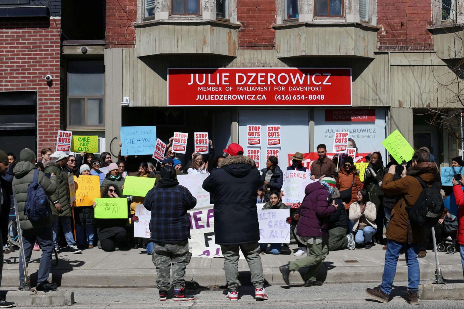 People are holding placards on the sidewalk outside the constituency office of Davenport MP Julie Dzerowicz. They are dressed in winter clothing and casual streetwear on a bright day.