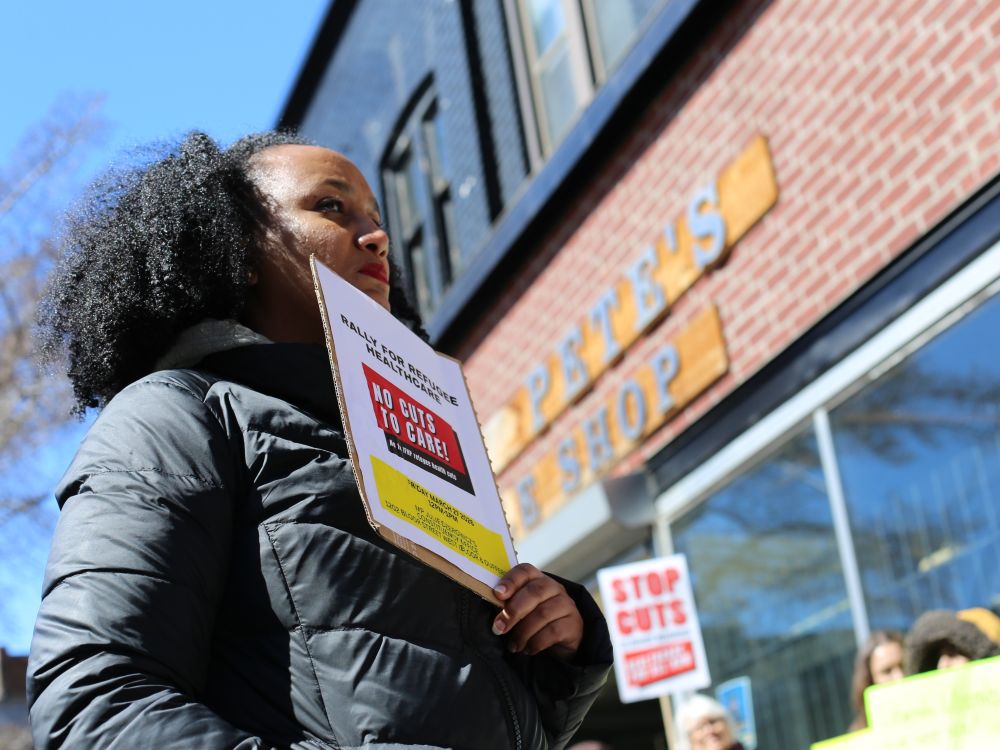 A woman with black curly hair is wearing a puffy black winter coat. She is holding a handmade sign that says, &ldquo;No cuts to care!&rdquo; She is standing outdoors outside a brick building on a clear day.