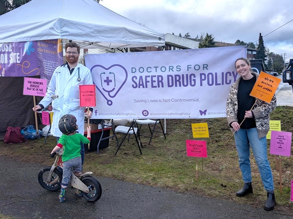 Two people stand on either side of a banner that reads, &lsquo;Doctors for Safer Drug Policy.&rsquo; In front of them is a child on a push bike.