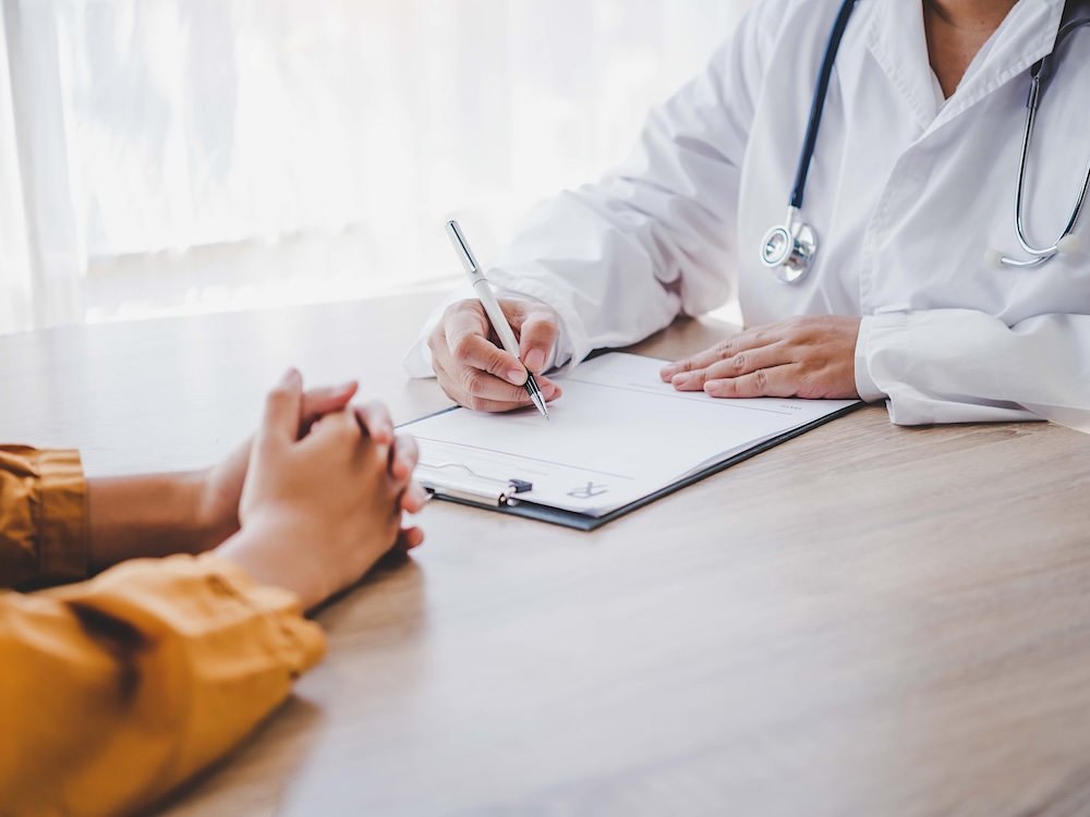 Two people sit at a desk, with only part of them visible. One is a doctor, wearing scrubs and writing on a notepad.