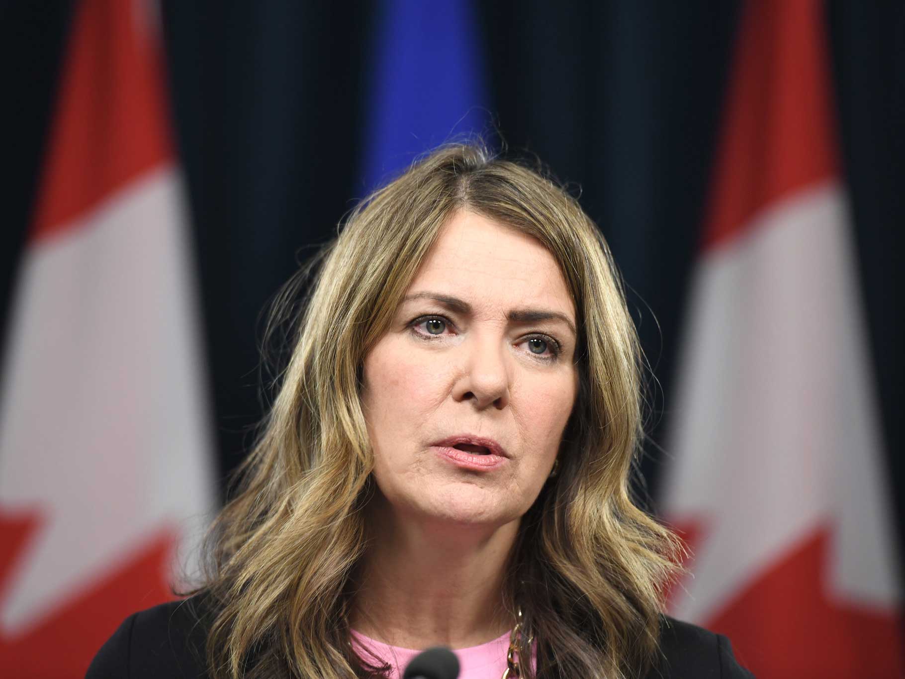 Danielle Smith, a 55-year-old woman with light-toned skin and shoulder length brownish hair stands at a podium. She wears a pink shirt and dark jacket.