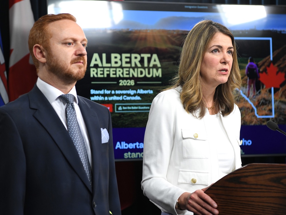 A 44-year-old man with reddish hair and wearing a blue suit stands beside Danielle Smith, who is wearing a white suit. Behind them is a monitor that says &ldquo;Alberta Referendum.&rdquo;