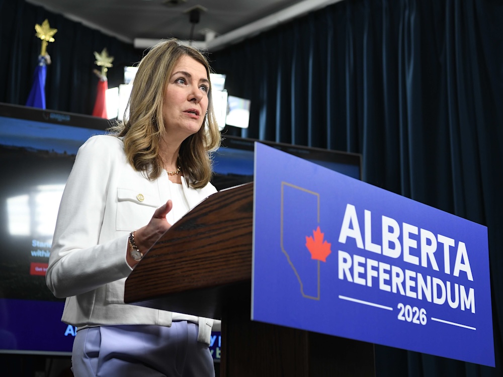Danielle Smith, a 55-year-old woman with light skin and brown shoulder-length hair, stands at a podium with a blue sign that says &lsquo;Alberta Referendum 2026.&rsquo; She wears a white suit.
