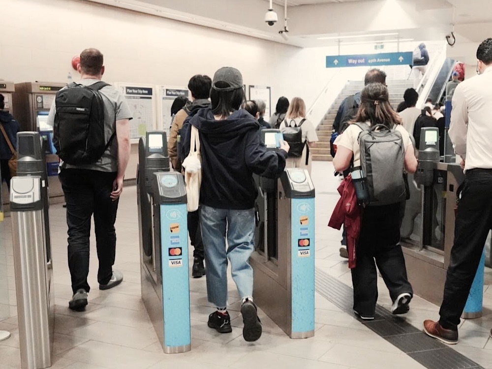 People in casual streetwear have their backs turned to the camera as they pass through blue and silver turnstiles in a public transit station. In front of them are white walls, a set of stairs and escalators.