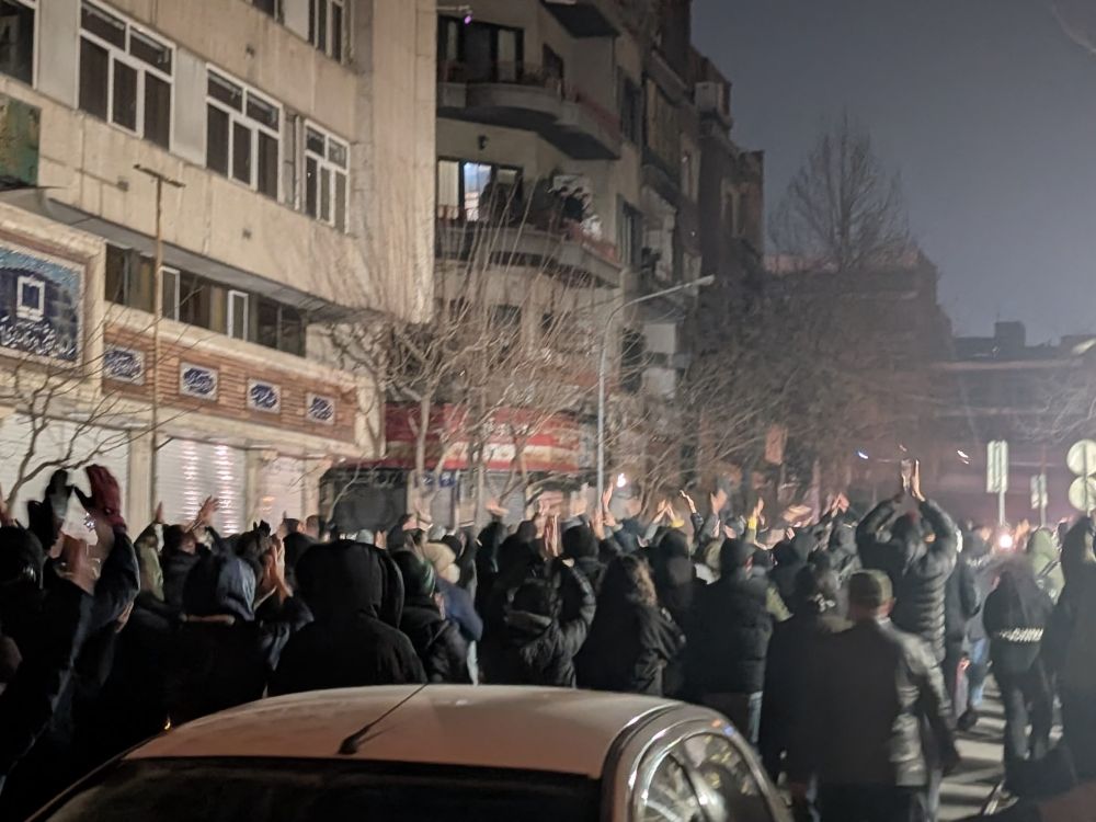 Protesters in dark clothing fill a street in Tehran at night, many with their arms raised. Their backs are turned to the camera.