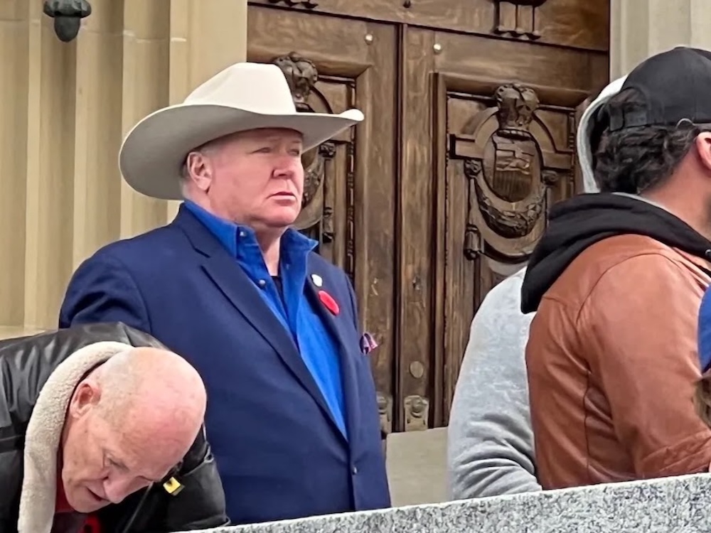 A burly late-middle age man in a white cowboy hat stands outdoors.