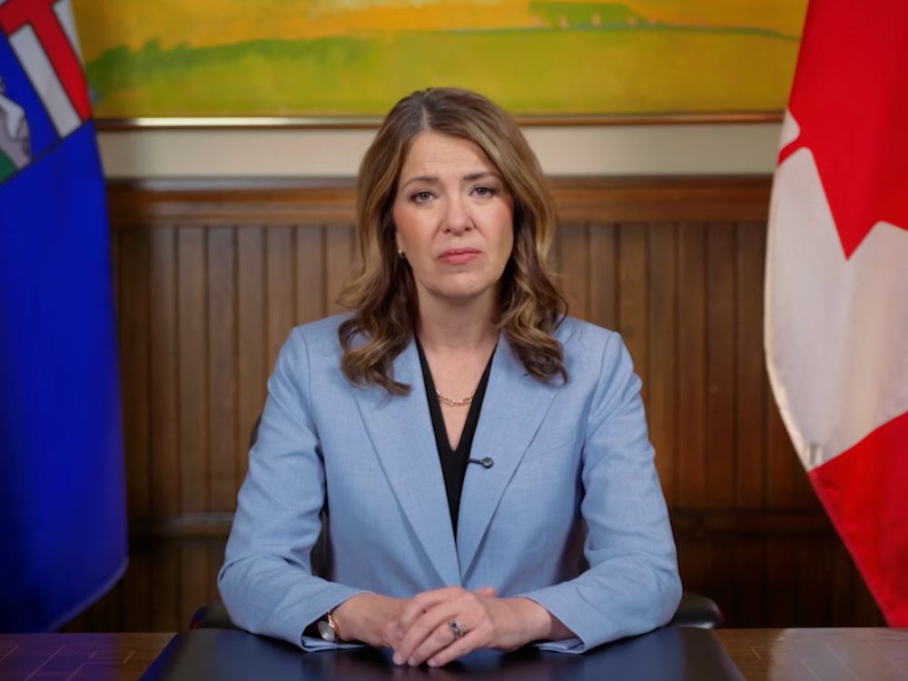Danielle Smith, a 55-year-old woman in a black shirt and light-blue suit, sits in front of flags and wood paneling with her clasped hands resting on a desk.