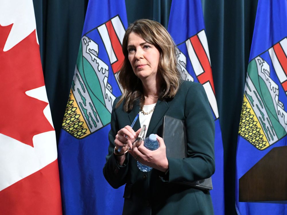 Danielle Smith, a 55-year-old woman wearing a green blazer and holding a water bottle, walks on to a podium. 