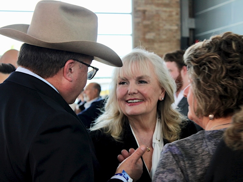 A 71-year-old woman with blond hair and light-toned skin smiles at a man in a light cowboy hat and a woman.