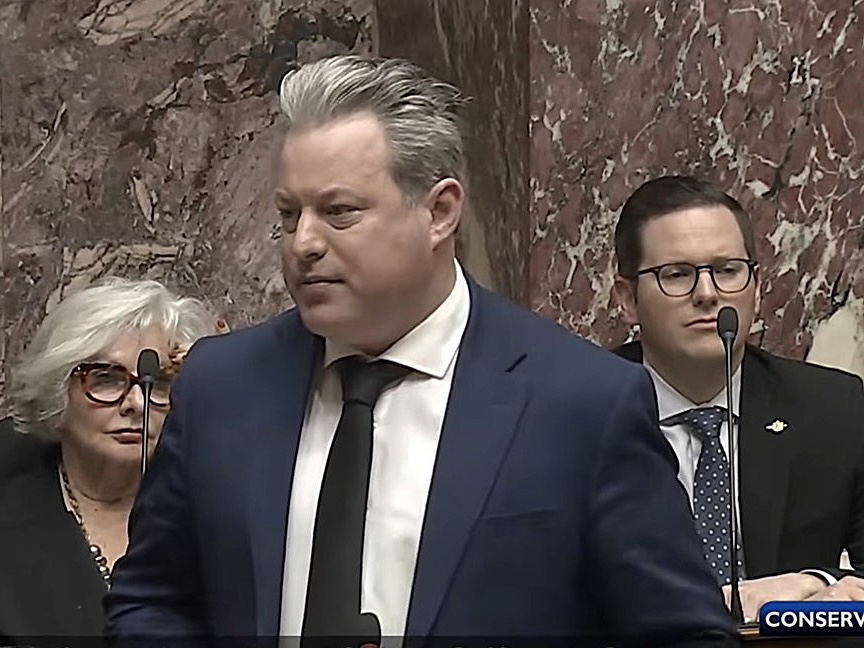 Trevor Halford, a man with light skin tone and grey hair, wears a suit and addresses the legislature.