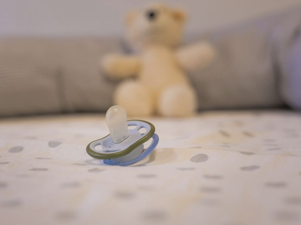 A baby pacifier on a blanket with a teddy bear in the background.
