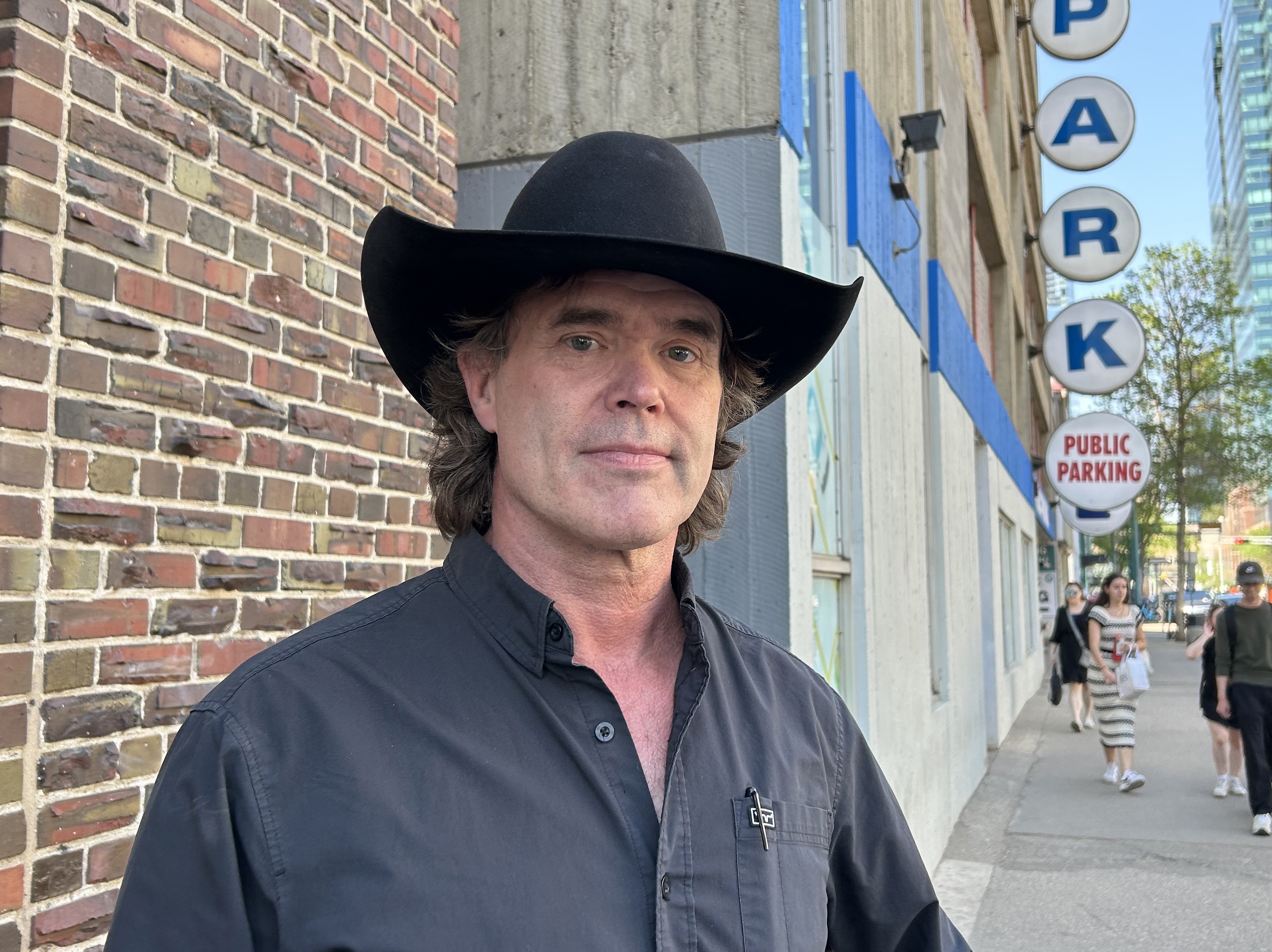A medium closeup shot of Corb Lund wearing a dark blue button-up and a black cowboy hat. He stands on a sidewalk subtly smiling into the camera. 