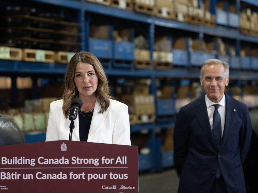 Danielle Smith, wearing a white jacket, stands at a podium in a warehouse with Mark Carney to the right.