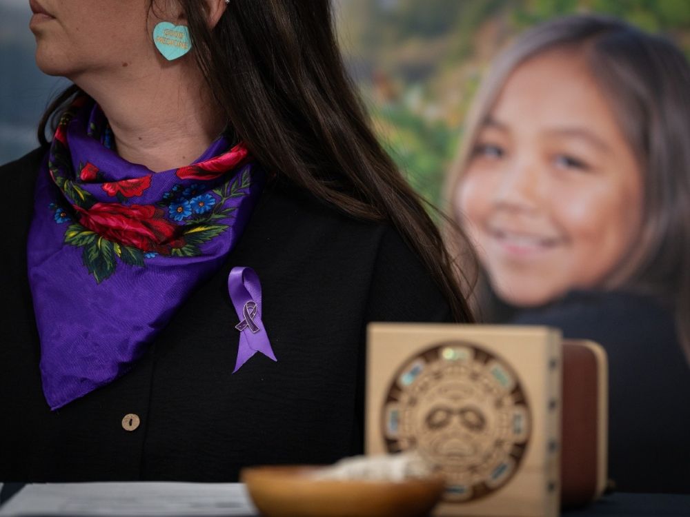 A purple ribbon is in focus on the lapel of a woman. Her eyes are obscured. In the background is an image of a young person looking at the camera. In the foreground, also blurred, is a piece of First Nations art.
