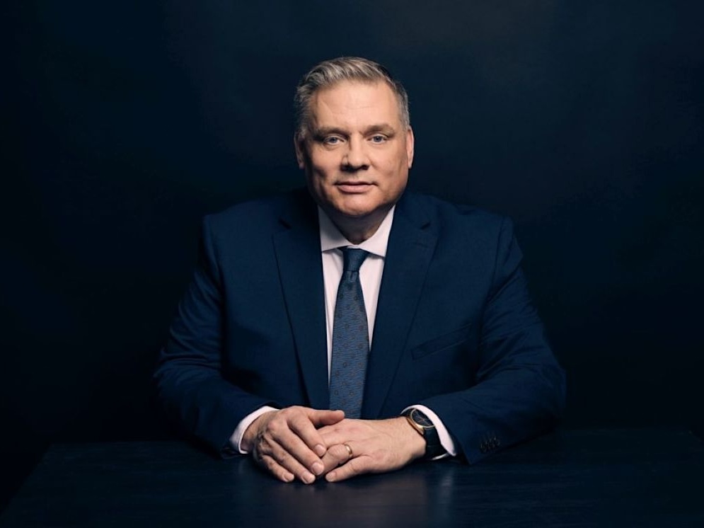 A middle-aged man with light skin and short ash-blond hair, wearing a dark suit and tie, sits with his hands in front of him.