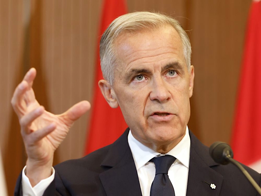 Mark Carney, a white man with short grey hair and wearing a dark suit and tie, gestures with one hand while speaking into a microphone. 