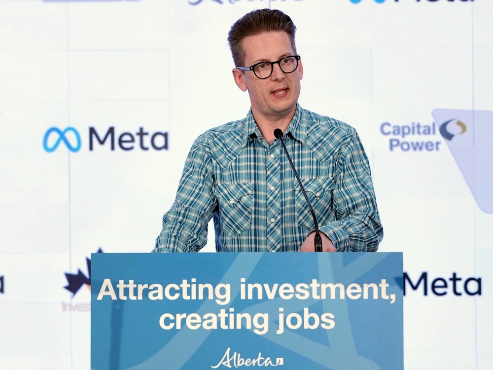 A 45-year-old man with light-toned skin, short hair and glasses, wearing a blue plaid western shirt, stands at a podium with a sign that says &lsquo;Attracting investment, creating jobs.&rsquo;