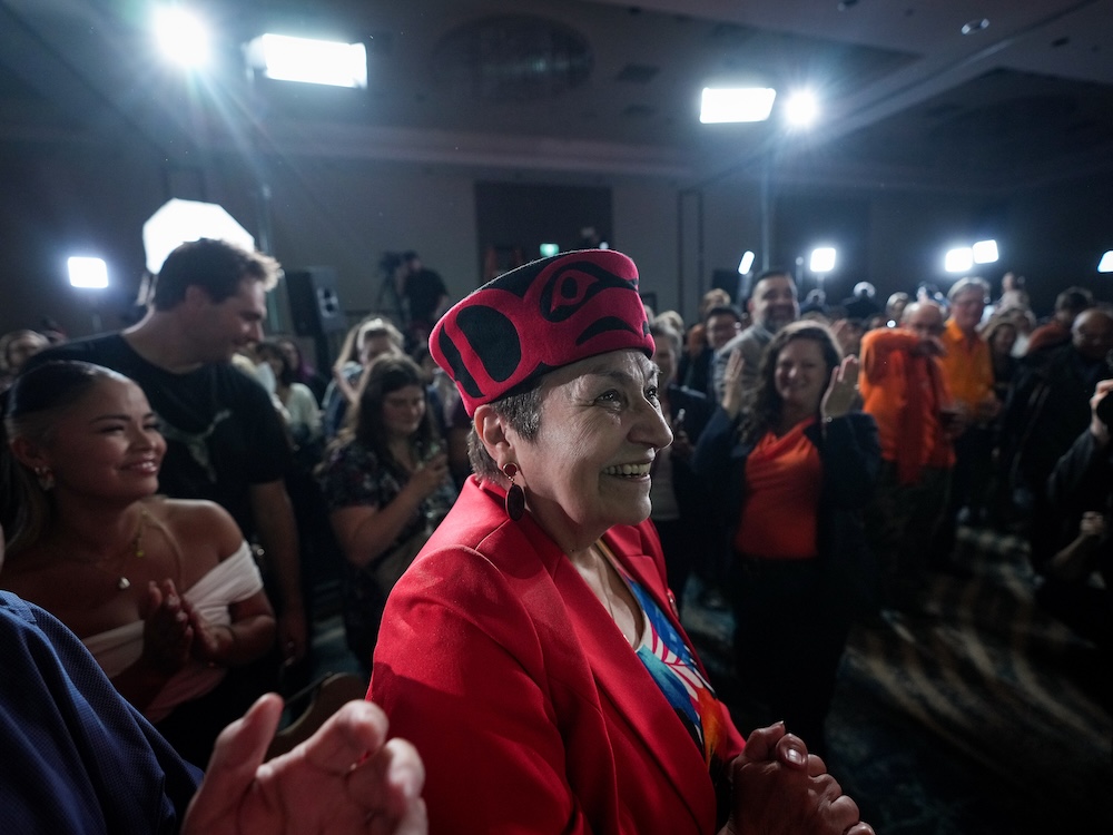 A 70-ish Indigenous woman wearing a red hat and jacket smiles, surrounded by supporters.