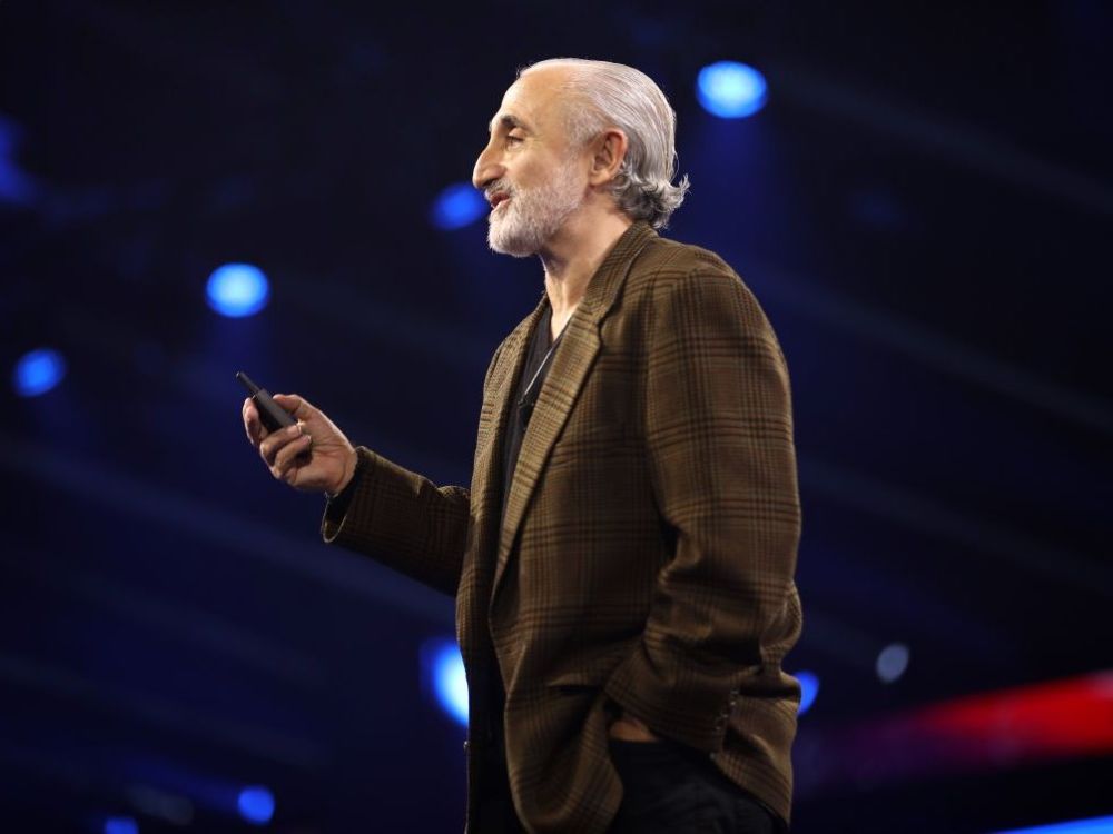 A man with a grey beard and slicked-back grey hair speaks from a stage.