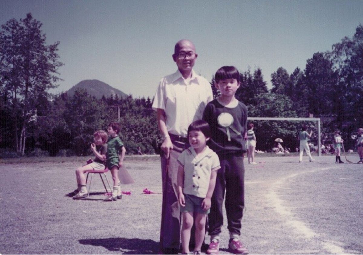 A man and two children stand on a fielded area in a park in an older colour photo.