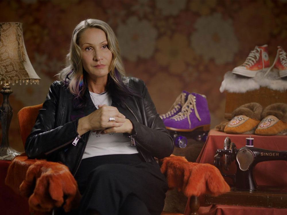 A woman sits in an orange armchair surrounded by vintage artifacts: an old Singer sewing machine, a pair of purple rollerskates, an art nouveau lamp and a pair of beaded moccasins.