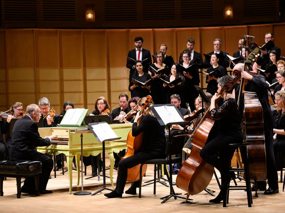 A man in a suit sits infront of a canary yellow grand piano, surrounded by a string orchestra and choir. There is a wooden backdrop and theatre lighting.