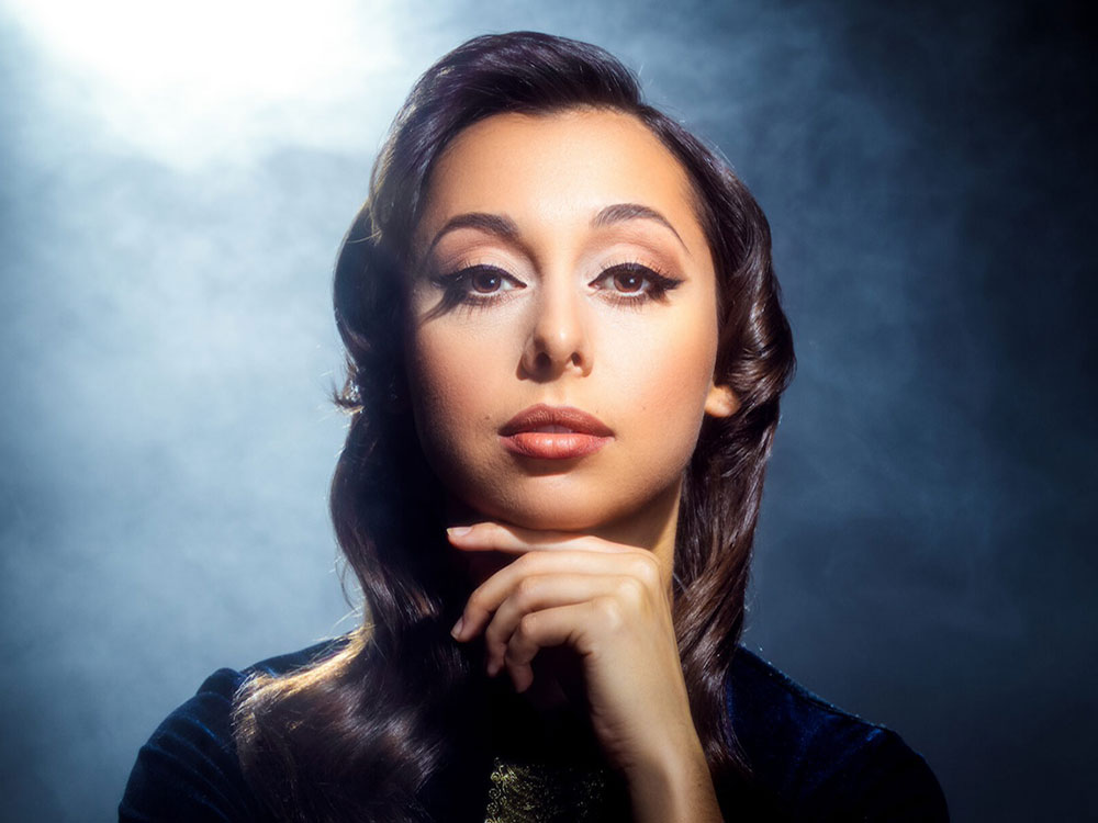 A woman looking directly at the camera rests her face on her chin amidst a blue-tinged smoky backdrop.

