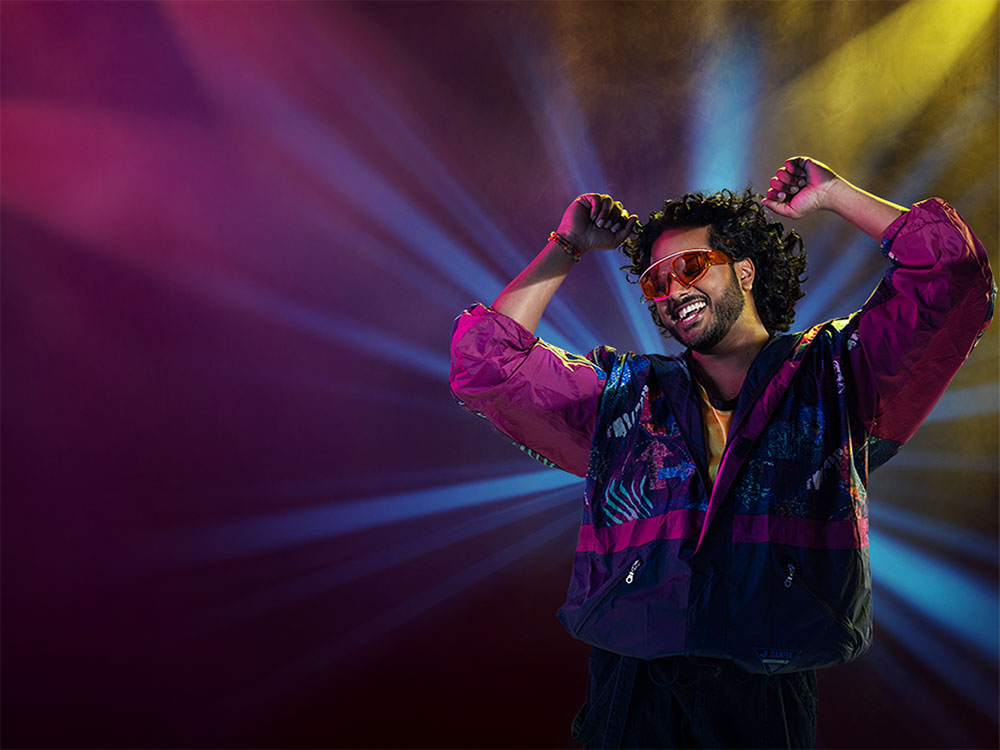 A person with black shaggy hair and beard wears a flashy track jacket and orange sunglasses. They are dancing in front of spotlights that accent their jacket.
