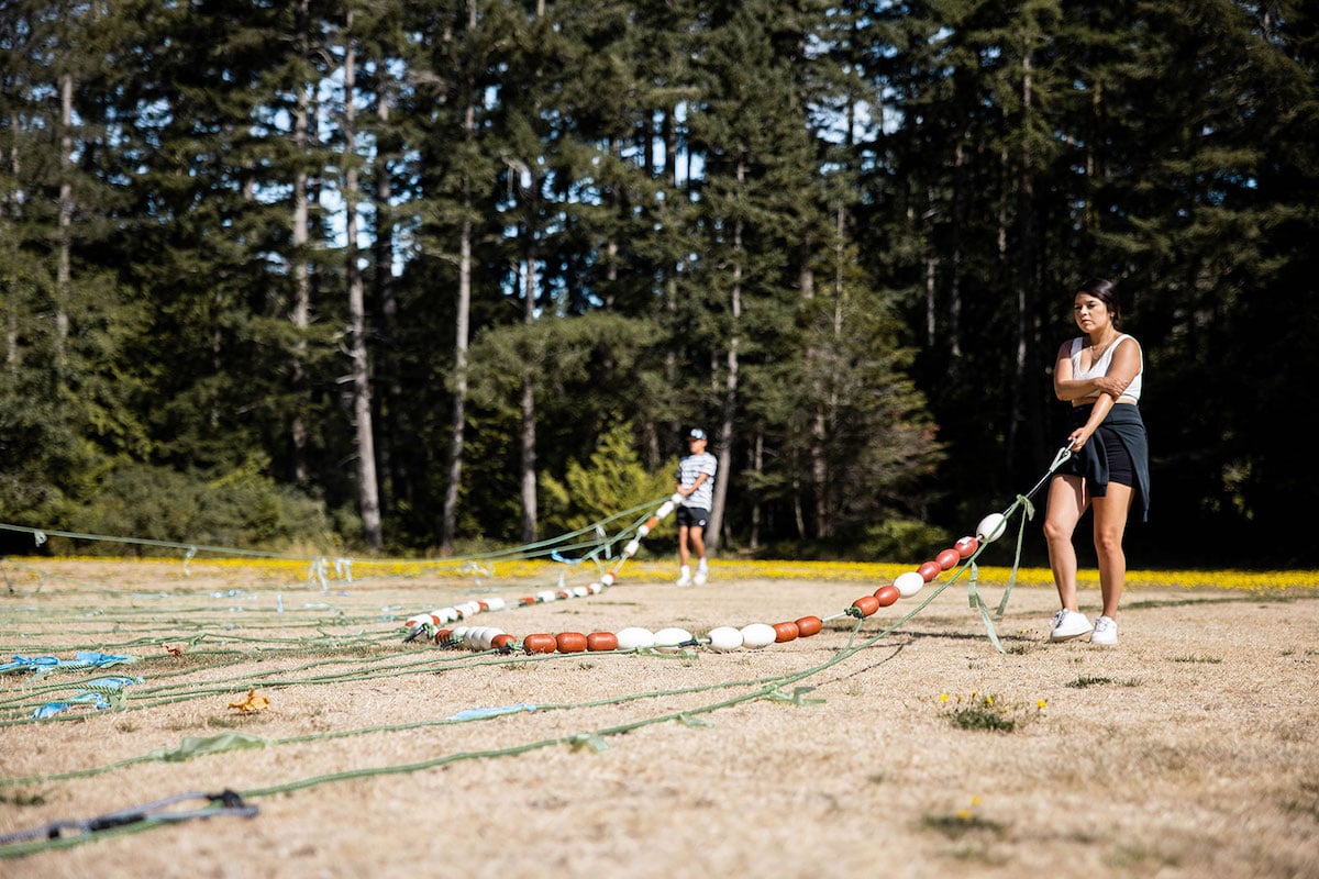 Two youth, one in the foreground and one in the background, pull ropes with buoys across the beach.