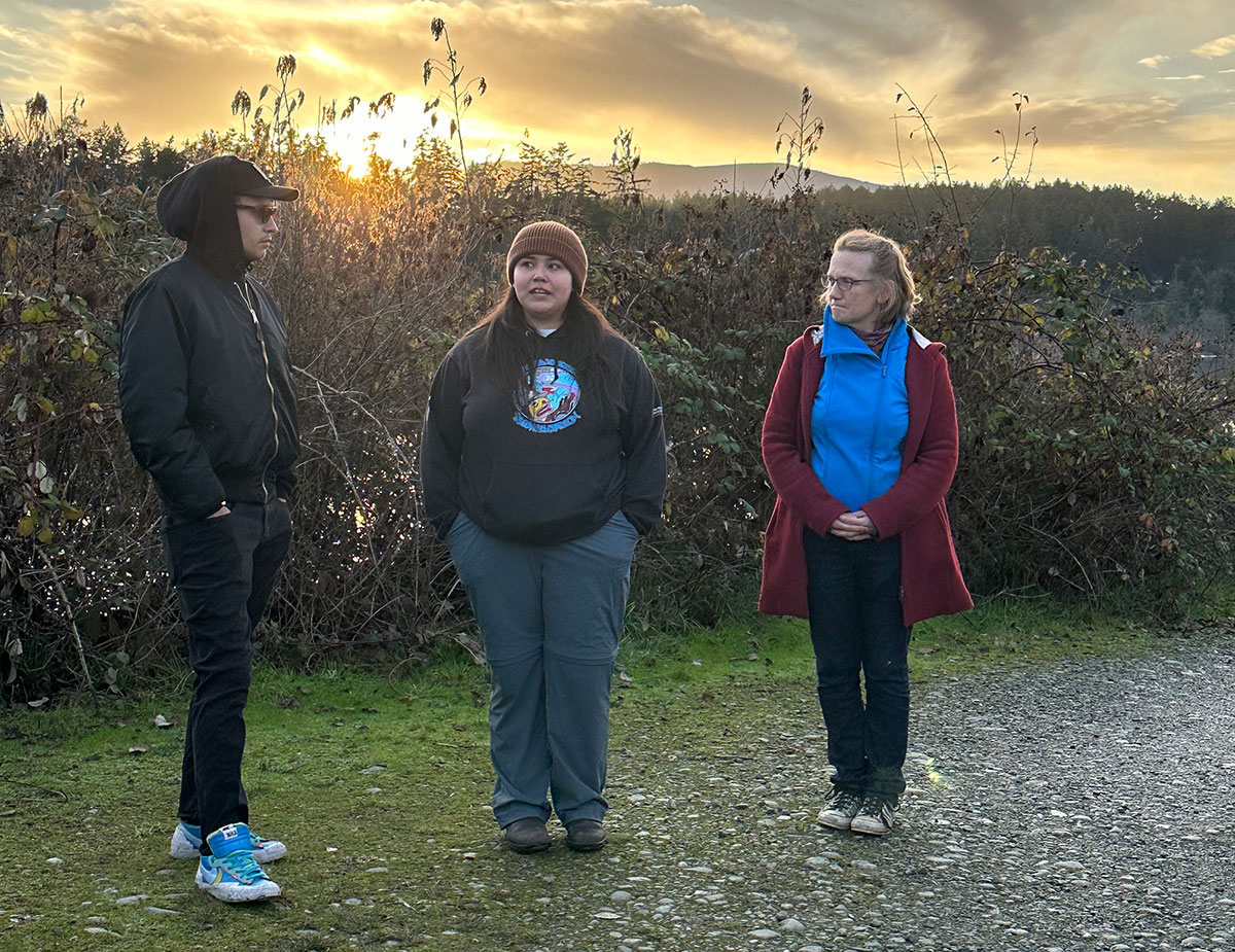 A woman with medium skin in a black hoodie, brown toque and grey pants stands with two other people in a clearing. The sun is dropping in the background behind them.