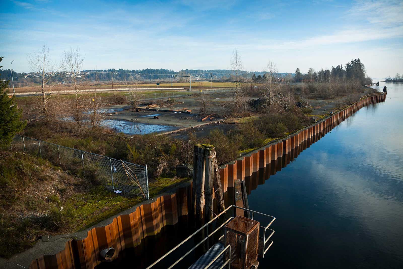 An aerial photo shows an abandoned sawmill site surrounded by a metal fence on two sides and a river on the other. The river is separated by a metal piling wall.