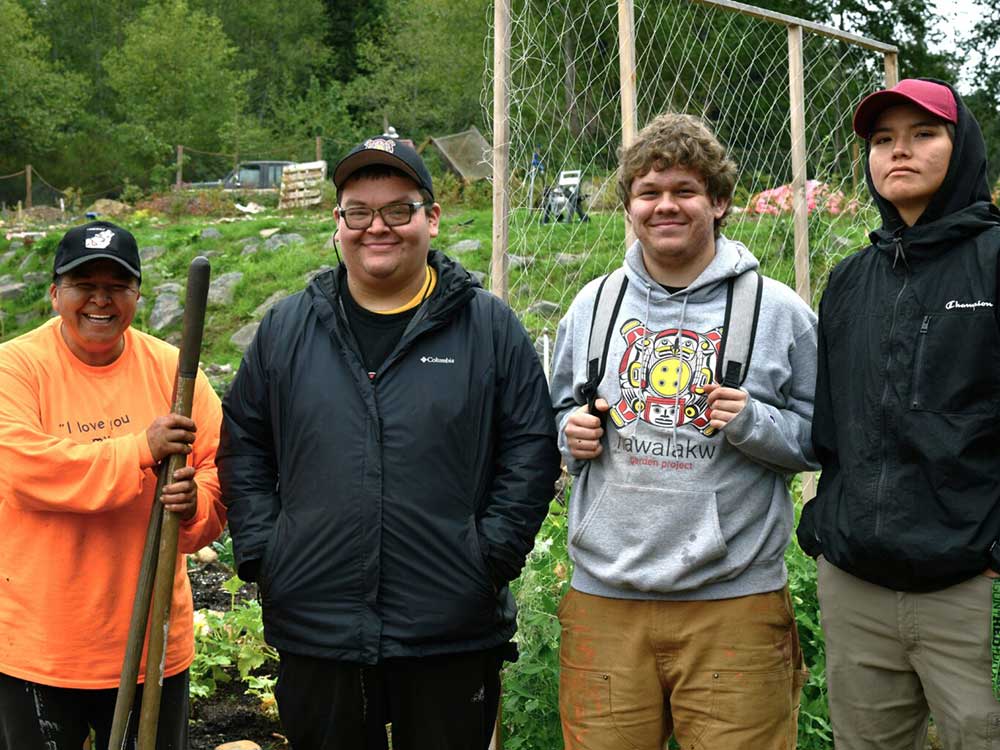 A group of four people stand in front of a thriving garden. At left, a person in an orange shirt is smiling and holding gardening tools. Three youth stand to her right.