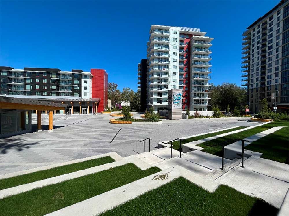 A concrete plaza with tiered grass landscaping is in the foreground of a residential tower and longer, five-storey building.