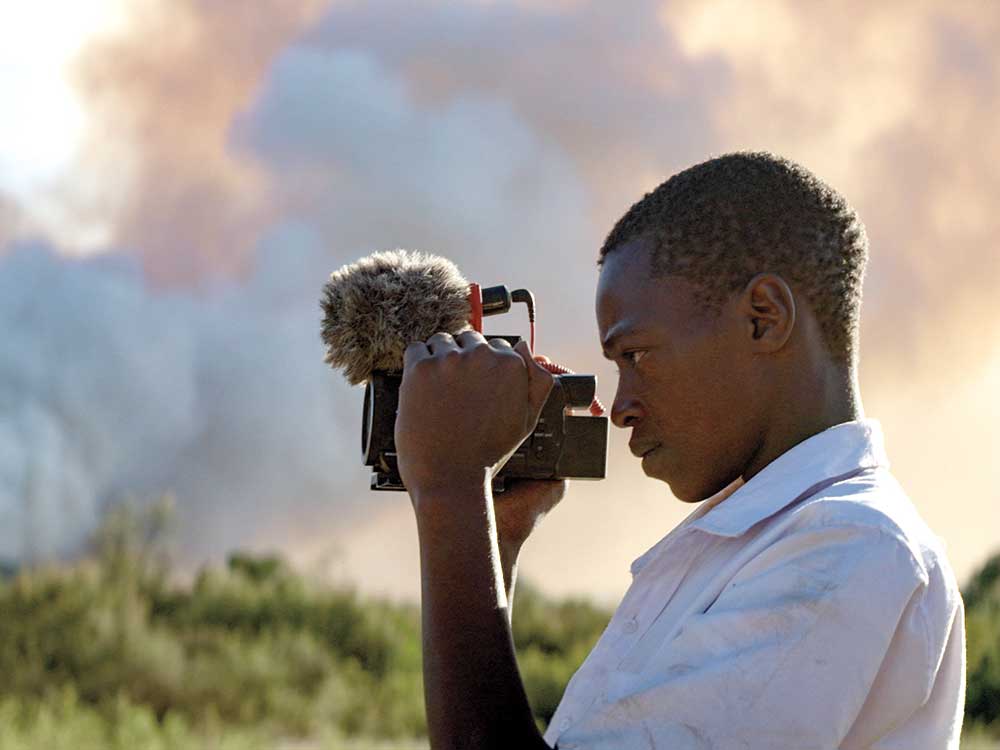 A boy with dark skin holds a personal video camera and looks into the lens. He is wearing a white collared shirt and standing in a clouded grass area.