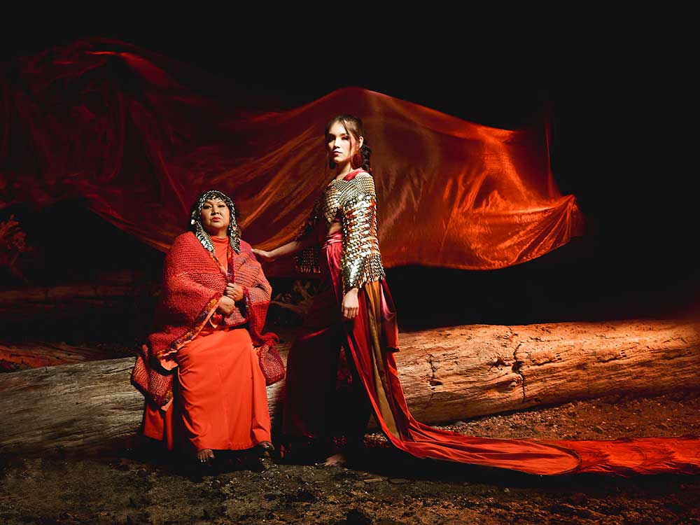 Two women are on a stage. One sits. One stands. They are dressed in red fabrics with metal accents. In the background, there is a big piece of flowing red fabric.