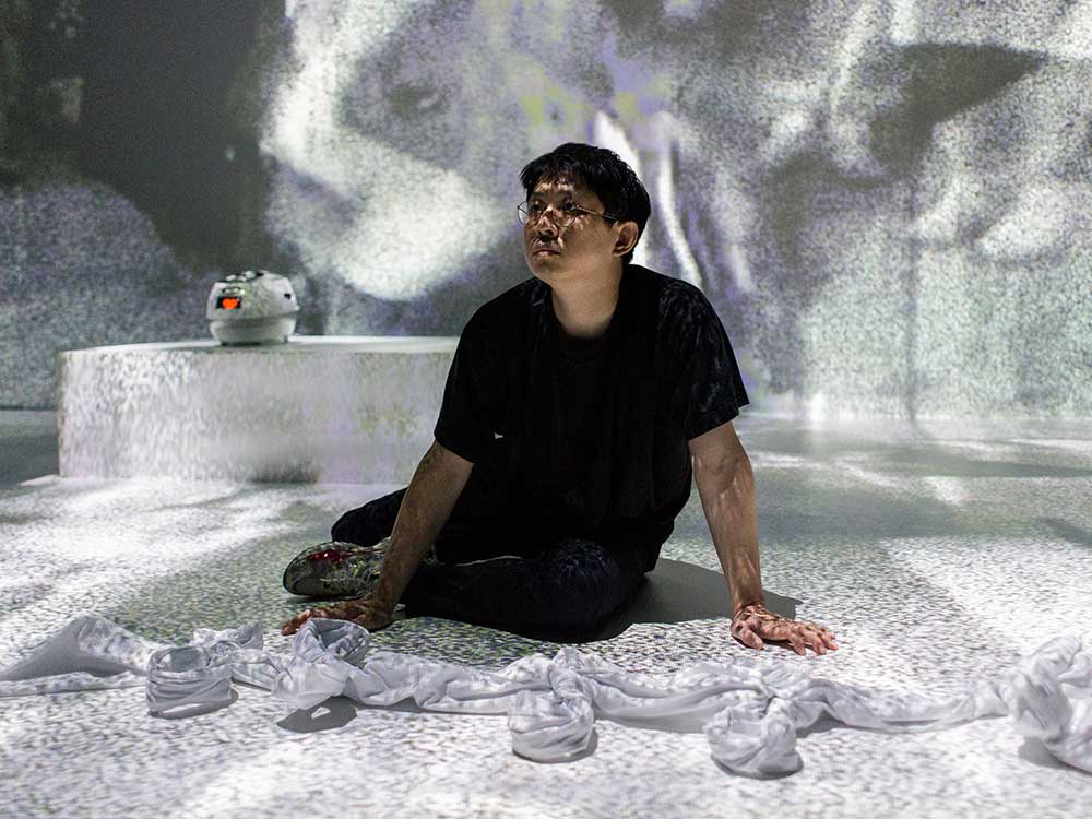 A person with medium-skin and black hair sits on a white stage, gazing forward. They are wearing a black T-shirt and black pants. Projections cover the stage.