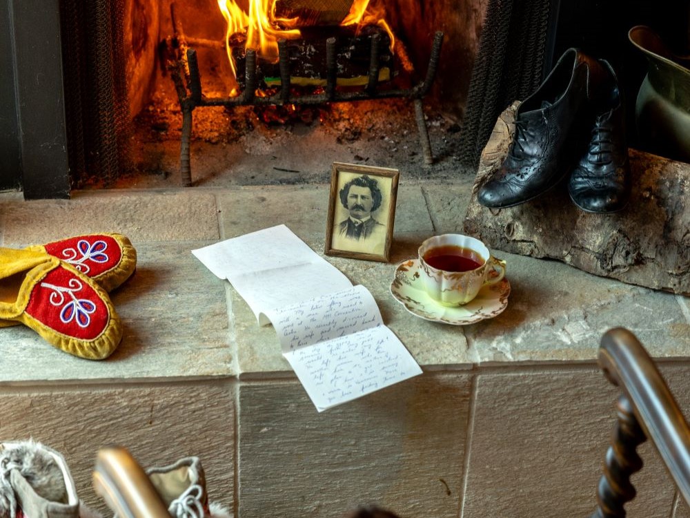 A fire burns on a hearth. On the hearth is a handwritten letter, a framed black and white pencil drawing of Louis Riel, a tea cup, a pair of mockasins and a pair of black leather lace-up shoes.
