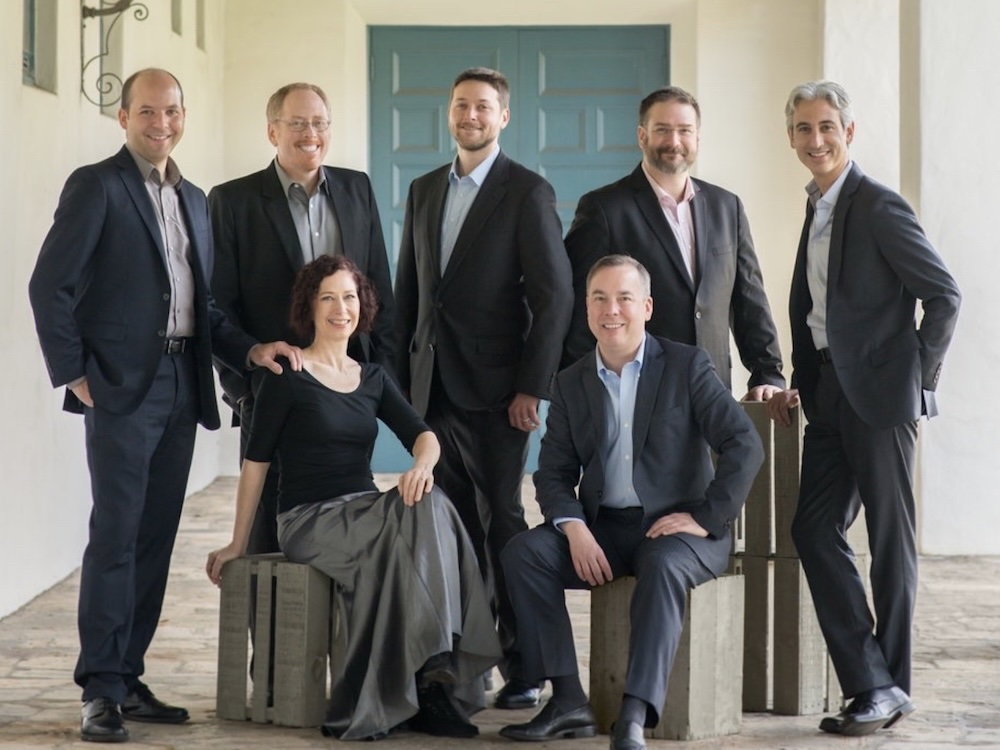 An ensemble consisting of six men in dark-coloured suits and one woman in a black blouse and dark grey skirt. Five of the men are standing while the woman and one man are seated on crates in front.