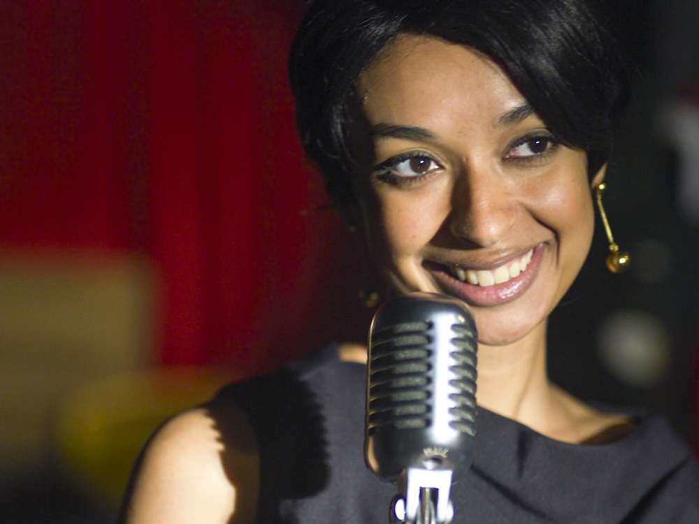 A woman with dark hair in a 1950s-style updo stands before a microphone onstage, smiling. The background is red and gold in soft focus.