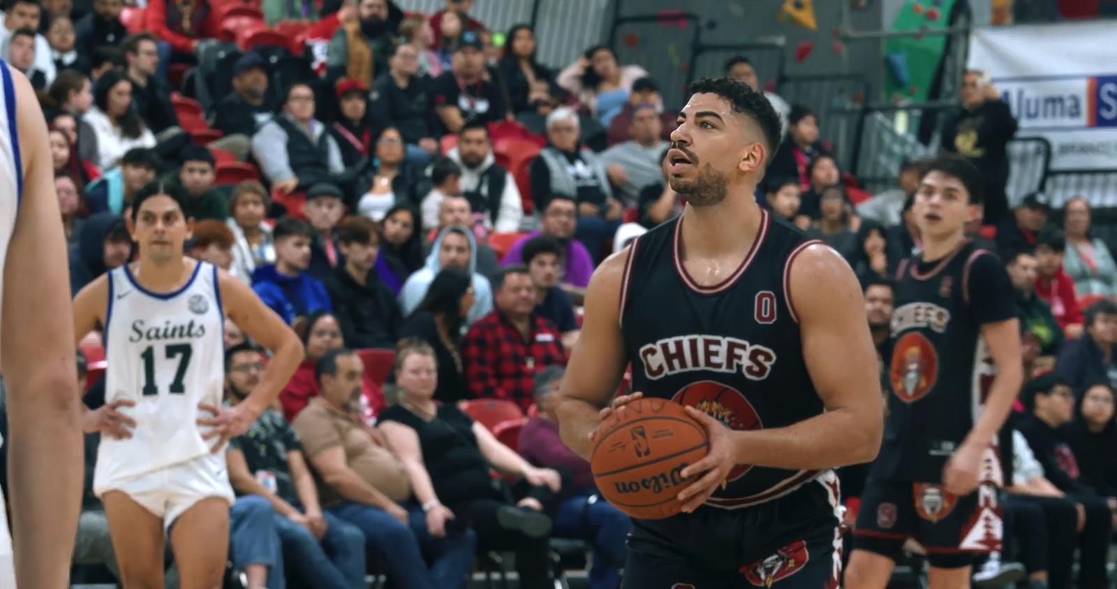 A young man with short dark curly hair, facial hair and a navy and red basketball uniform stands in the middle of a gymnasium, holding a basketball. Behind him are other players and red seats filled with people.