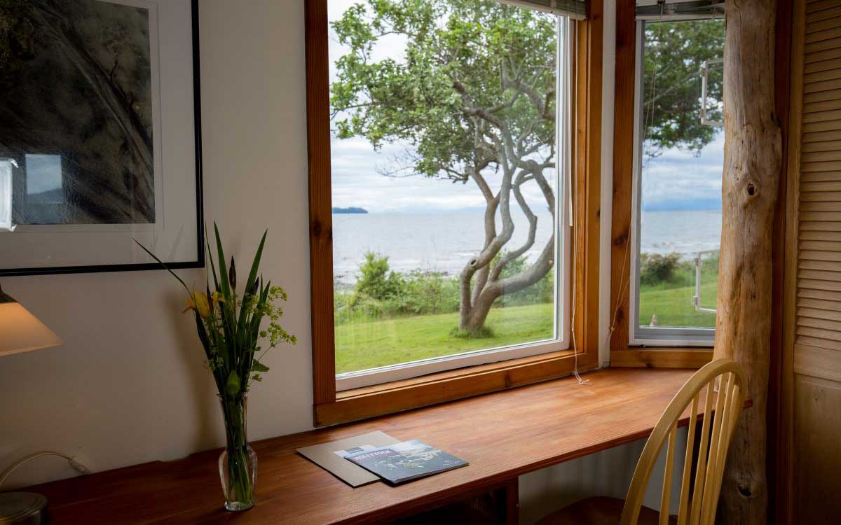 A wooden desk adorned with flower vase sits in front of a window with a bright view of a lawn, tree and a body of water.