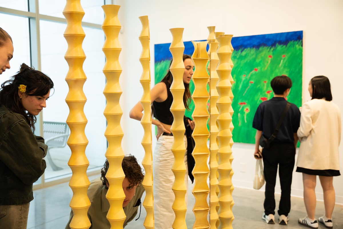 Three pairs of students stand in an indoor gallery space interacting with the visual art on display. In the foreground is an installation featuring tall yellow pillars in an exaggerated shape.