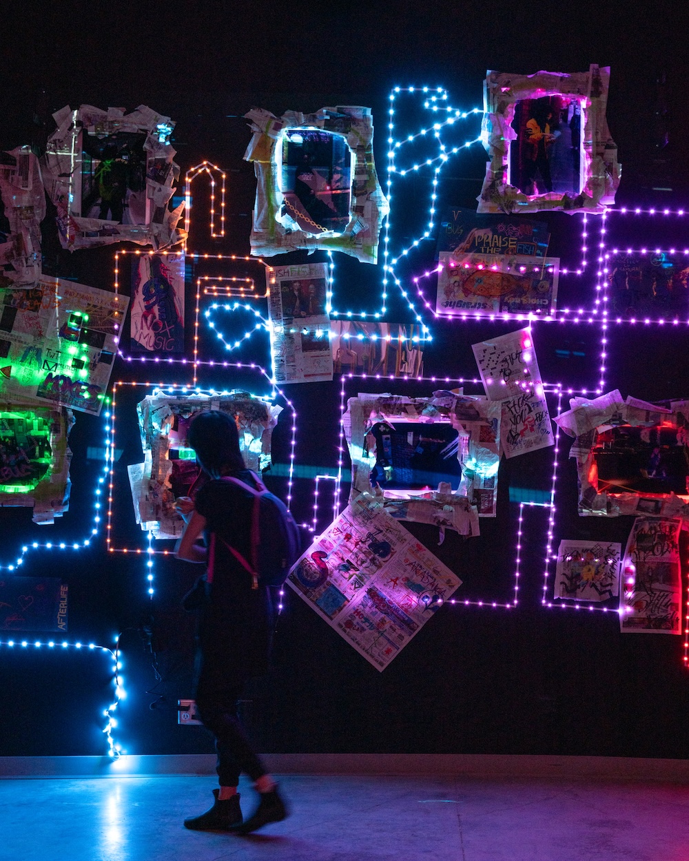 A person with dark hair, a T-shirt and a backpack is stepping towards a large-format art installation that features a geometric pattern of colourful lights against mixed-media artwork, including newspapers and print ephemera, mounted to the wall in a dark gallery space.