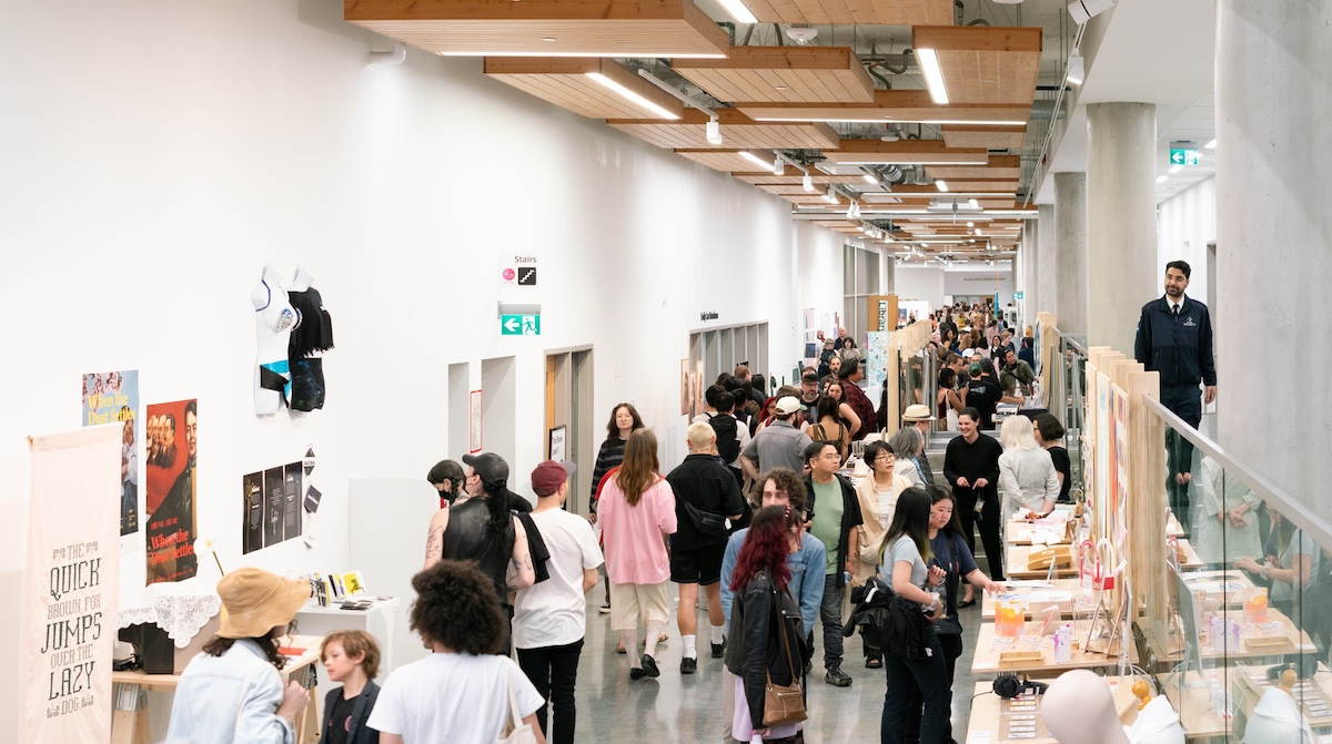 Large groups of people are walking, mingling and observing art in a crowded concourse in an indoor space with wood beams on the ceiling and white walls. Artwork is displayed on tables to the right of the frame as well as alongside the left walls.
